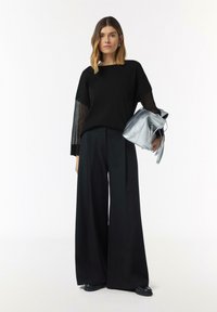 Schwarzer Pullover mit durchsichtigen Ärmeln, kombiniert mit weiten schwarzen Hosen. Hält eine silberne Handtasche. Glatte Textur und minimalistisches Design.