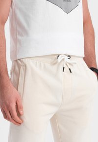 Beige Baumwoll-Jogginghosen mit elastischem Bund, Kordelzug und seitlichen Taschen. Kombiniert mit einem weißen Shirt mit geometrischem schwarzen Druck.