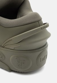 Ελαιόχρωμο αθλητικό παπούτσι με λείο δερμάτινο σώμα, υφασμάτινη περιφέρεια με υφή και έντονη κυκλική σφραγίδα στην σόλα. Χαμηλού προφίλ σχέδιο.