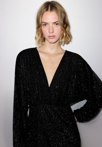 Blonde Frau mit kurzen welligen Haaren, die ein schwarzes, glitzerndes, tief ausgeschnittenes Kleid trägt und mit einer Hand in der Hüfte vor einem einfarbigen Hintergrund steht.