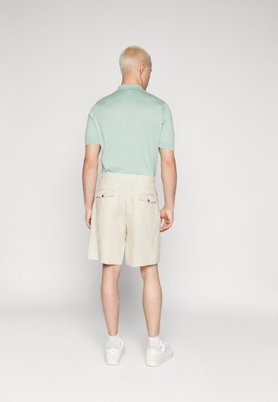 Polo shirt vert menthe à manches courtes, associé à un short beige avec poches arrière et accents en boutons. Porté avec des baskets blanches.