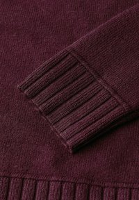 Detail eines burgunderfarbenen Strickpullovers, der einen gerippten Bund und Saum mit einem strukturierten Muster zeigt. Das Material wirkt weich und warm.