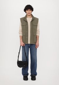 Paceco verde oliva senza maniche con più tasche sopra un maglione beige, abbinato a jeans a gamba larga blu e una borsa a tracolla nera.