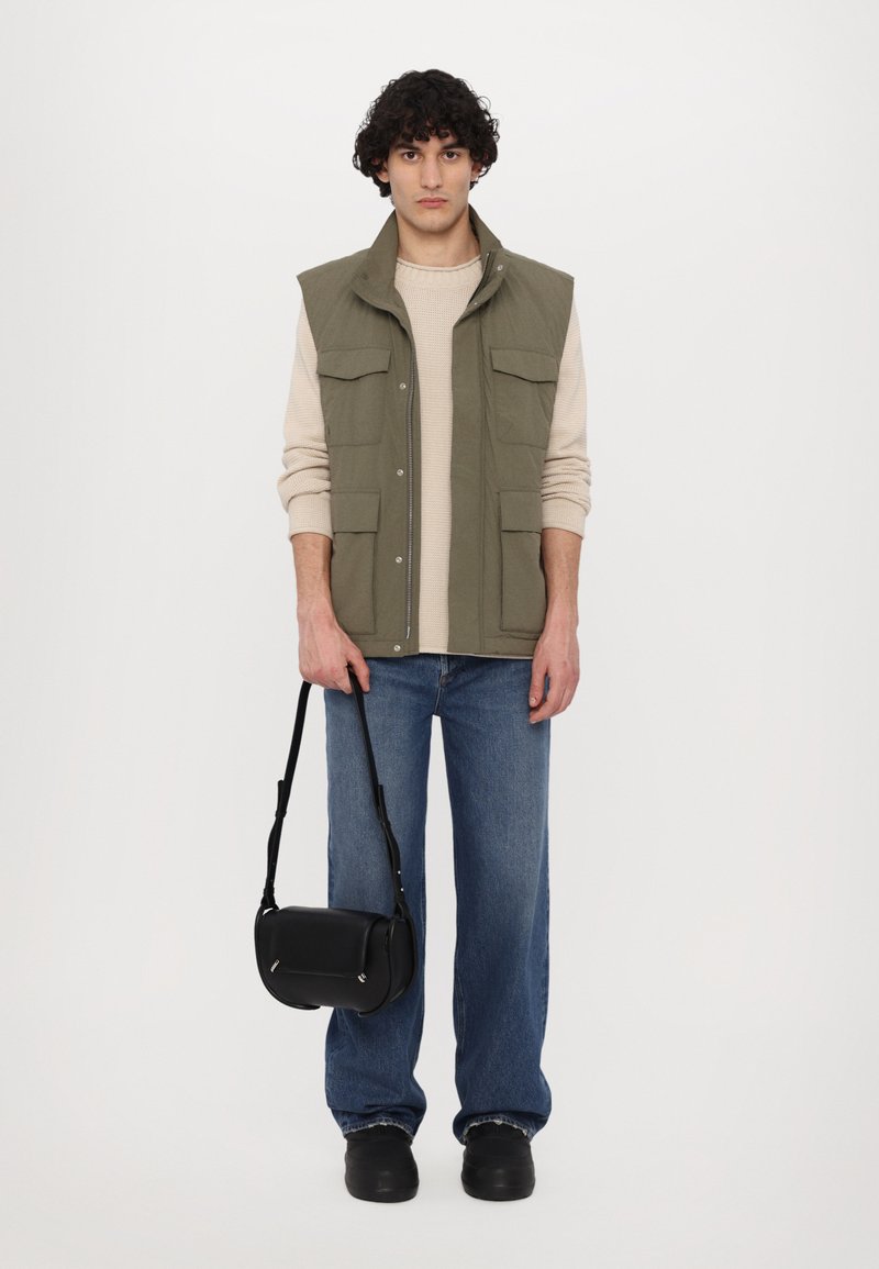 Paceco verde oliva senza maniche con più tasche sopra un maglione beige, abbinato a jeans a gamba larga blu e una borsa a tracolla nera.