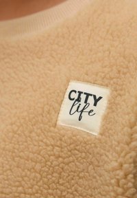Tan fleecová látka se strukturovaným povrchem a bílým žebrovaným výstřihem. Čtvercová visačka obsahuje slovo "CITY" tučným černým písmem a "life" v kurzívě.