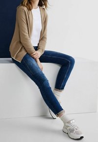 En tanfärgad kofta över en vit skjorta, tillsammans med blå denimjeans. Ljusfärgade sneakers med reflekterande detaljer och ljusa strumpor fulländar outfitten.