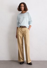 Kobieta z kręconymi włosami w jasnoniebieskiej bluzie Marc O’Polo, beżowych szerokich spodniach, brązowym pasku i ciemnych butach, stojąca przy białej ścianie.