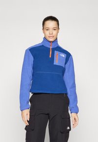 Fleece pulover u nijansama plave boje s visokim ovratnikom, četvrtastim zatvaračem i džepom s krpom. Sadrži narančasti zatvarač i detalje s logom.