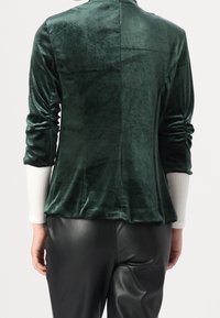 Grön sammetjacka med en strukturerad design, som har rynkade ärmar och hög krage, i kombination med svarta läderbyxor.