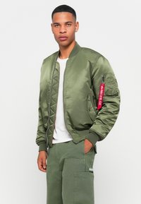 Man wearing olive green bomber jacket with red tag on sleeve, white shirt, and olive cargo pants, standing against a white background.