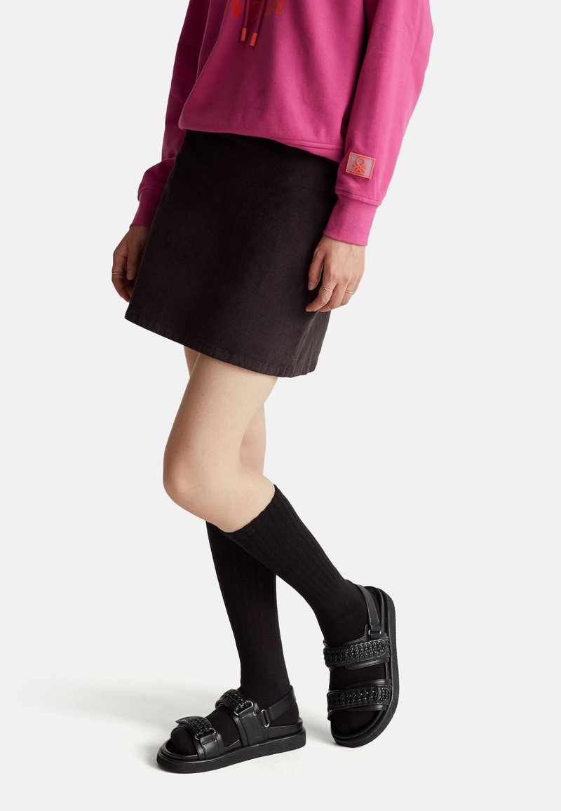 Černá kordurová mini sukně spárovaná s růžovou mikinou se oranžovými grafikami, žebrovanými černými podkolenkami a masivními černými sandály.