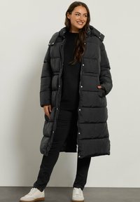 Abrigo puffer negro largo con capucha, que presenta un acolchado horizontal, cierres de botones y cremallera, y bolsillos laterales hechos de un material suave y mate.