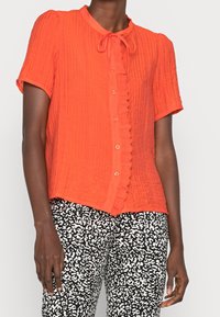 Short-sleeve, button-up blouse in bright orange textured fabric with a collar and a decorative ruffle along the front opening.