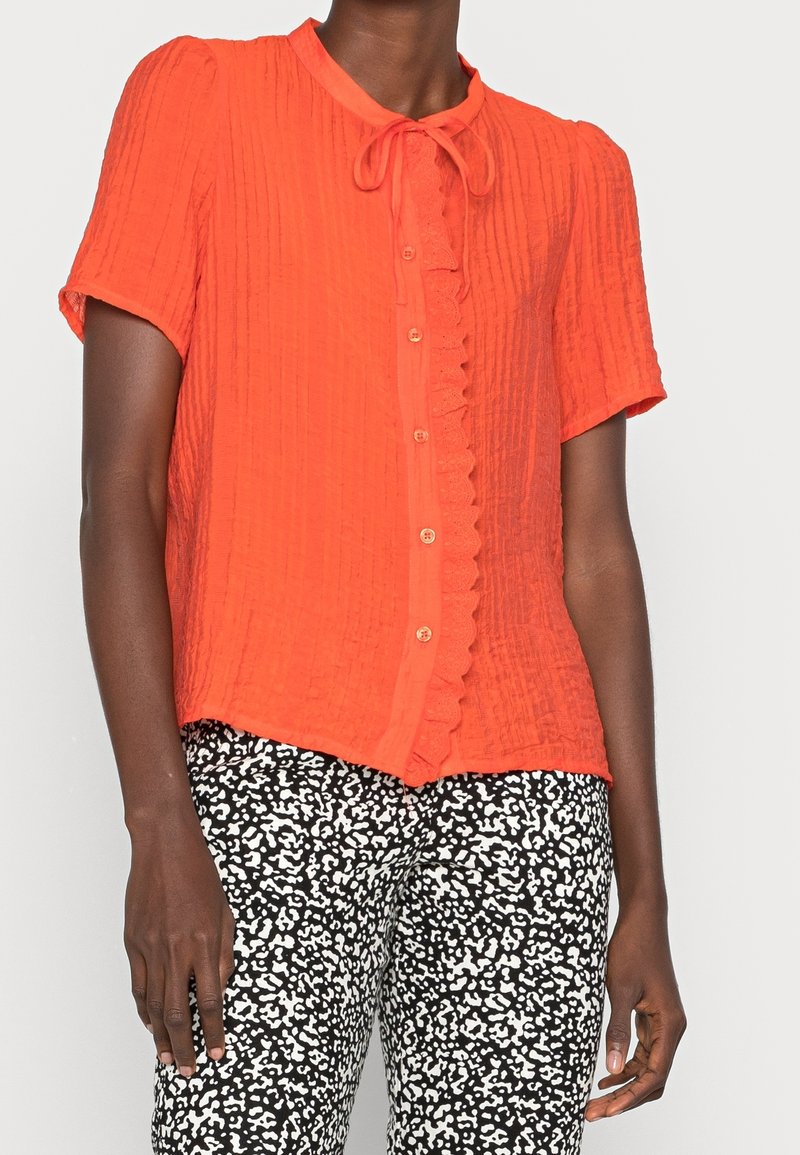 Short-sleeve, button-up blouse in bright orange textured fabric with a collar and a decorative ruffle along the front opening.