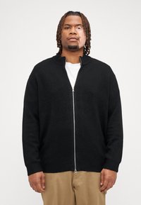 Sort lynlås cardigan med ribbet krave og manchetter, lavet af tekstureret stof, båret over en hvid T-shirt. Enkel, lige skåret design.