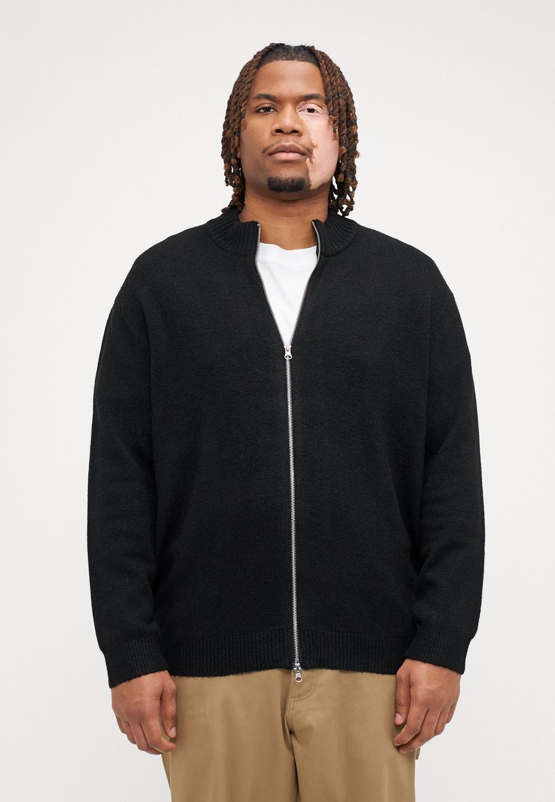 Sort lynlås cardigan med ribbet krave og manchetter, lavet af tekstureret stof, båret over en hvid T-shirt. Enkel, lige skåret design.