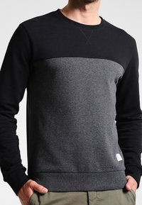 Sudadera negra y gris con cuello redondo, que presenta un diseño de bloques de color horizontal y puños y dobladillo acanalados. Textura de tela suave.