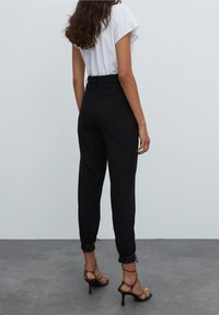 Pantalones negros de talle alto con piernas ajustadas, puños ceñidos y detalles de costura mínimos. Combinados con una blusa blanca y tacones destalonados.