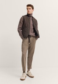 Lässige Outfit mit einer dunkelbraunen, gesteppten Weste, einem hellbraunen Strickpullover, beigen Hosen und weißen Sneakers mit tannenen Sohlen.
