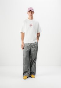 White oversized t-shirt with pink graphic print, gray shiny wide-leg trousers, and yellow shoes. Pink cap completes the outfit.
