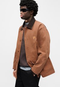 Καφέ τζιν μπουφάν με σκούρο γιακά, κλείσιμο με κουμπιά μπροστά και λογότυπο στο μπατζ. Φορεμένο πάνω από καφέ πουλόβερ και στρωμένο με ανοιχτό μπλε πουκάμισο.