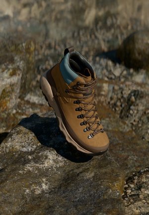 Botte de randonnée marron avec col de cheville rembourré bleu reposant sur une surface rocheuse rugueuse en plein air sous lumière naturelle.