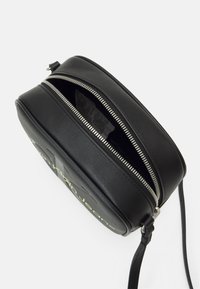 Kulatá černá kožená krosná taška se zipovým uzávěrem, s texturovaným povrchem a kontrastním žlutým logem na přední straně.