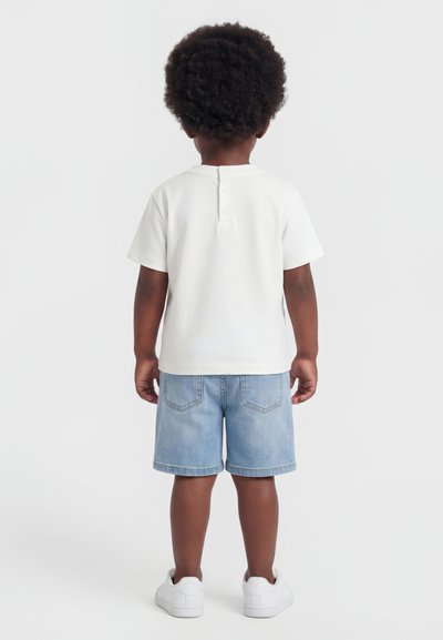 Παιδί με κοντά σγουρά μαλλιά που φορά λευκό κοντομάνικο πουκάμισο, ανοιχτό μπλε τζιν σορτς και λευκά αθλητικά παπούτσια, στέκεται γυρισμένο προς τα πίσω.