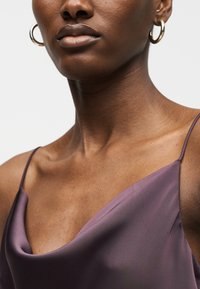 Κοντινό πλάνο ενός ατόμου που φοράει μωβ σατέν καμιζόλ και χρυσούς κρίκους σκουλαρίκια, με έμφαση στον λαιμό, τους ώμους και τα χείλη.