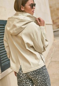 Béžová bunda s kapucňou a elastickými manžetami, s detailom ventilačky na chrbte, skombinovaná so sivými nohavicami so vzorom leopardích škvŕn. Textúrovaná a ľahká látka.