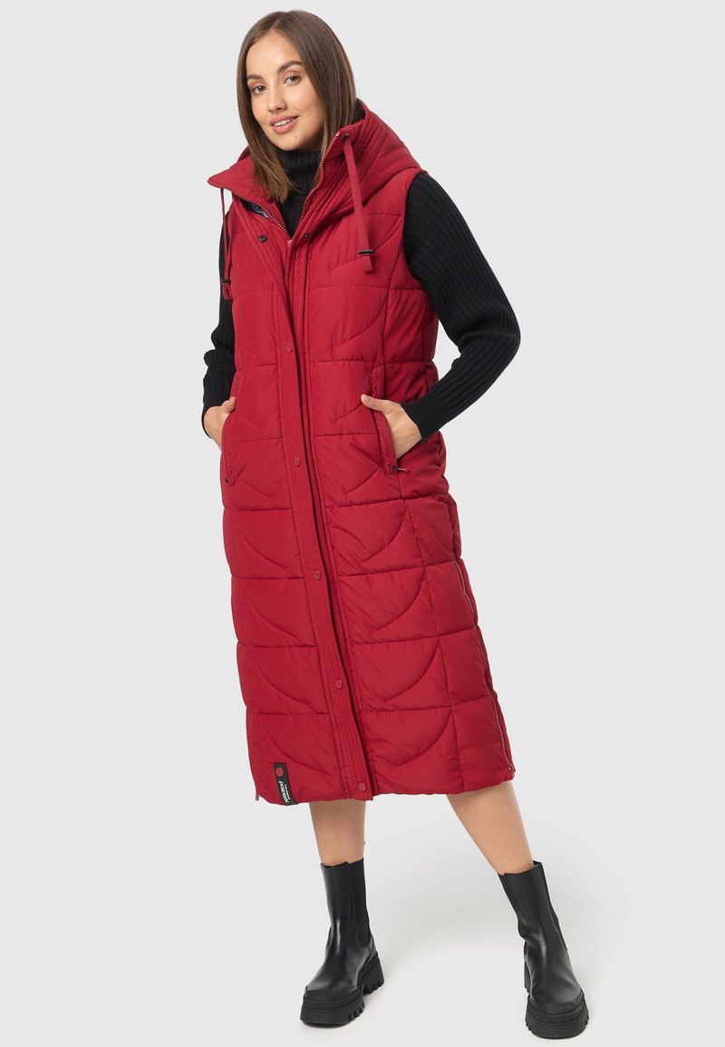 Longue veste rembourrée rouge avec col haut, fermeture éclair à l'avant et poches. Présente un design matelassé et est associée à des bottines noires à la cheville.