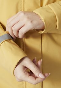 Chaqueta impermeable amarilla con un ajuste ceñido. La manga tiene un acento gris. Primer plano de manos ajustando la zona de la muñeca, mostrando la textura suave de la tela.