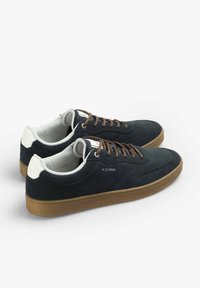 Zapatillas de ante azul marino con cordones marrones, detalles blancos en el talón y una suela de goma. Presenta un logo embellecido en el lateral.