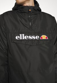 Giacca nera con cappuccio realizzata in tessuto liscio, presenta una tasca frontale prominente con il logo bianco "ellesse" e un accento colorato sotto il logo.