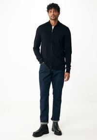 Svart zippered cardigan med hög krage, bärs över en ljus skjorta och matchas med mörkblå uppvikta jeans och svarta ankboots.