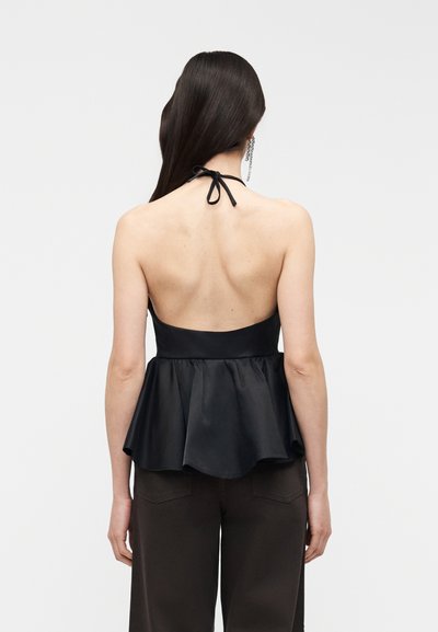 Femme aux longs cheveux foncés portant un haut à col licou noir avec un nœud au cou et une taille évasée, associé à un pantalon foncé, vue de dos sur un fond blanc.