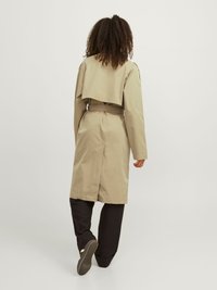 Beige Trenchcoat mit einem drapierten Rückendesign, tailliertes Modell und langen Ärmeln, getragen über schwarzen Hosen und Schuhen, vor einem schlichten Hintergrund.