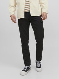 Black cargo pants with side pockets, paired with a beige button-up shirt and black sneakers with white laces. Textured fabric, casual fit.