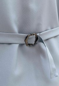 Lys grått stoffbelte festet med en mørk metallisk rektangulær spenne på en jevn matchende stoffbakgrunn.