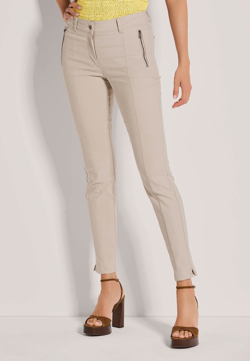 Beige Skinny-Hosen mit glatter Textur, Reißverschlusstaschen und seitlichen Details, kombiniert mit braunen High-Heel-Sandalen.
