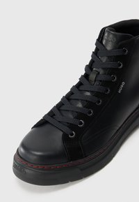 Zapatilla alta negra de cuero con un acabado texturizado. Presenta cordones negros, ojales plateados, costuras rojas y una suela de goma gruesa.