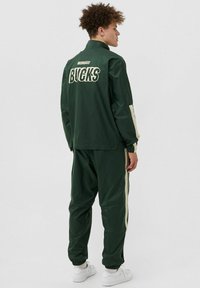 Completo sportivo verde realizzato in tessuto liscio. Dotato di un alto colletto, polsini elasticizzati e strisce laterali di colore chiaro. Logo "BUCKS" sul retro.