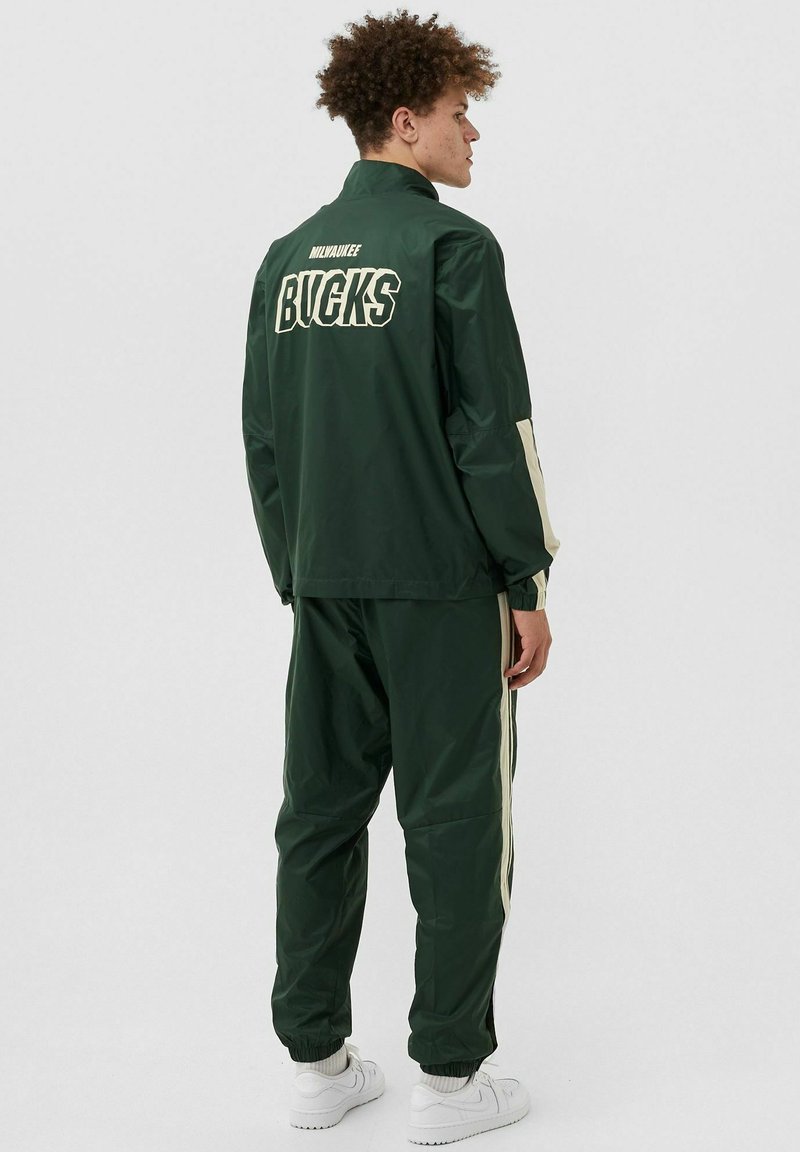 Completo sportivo verde realizzato in tessuto liscio. Dotato di un alto colletto, polsini elasticizzati e strisce laterali di colore chiaro. Logo "BUCKS" sul retro.