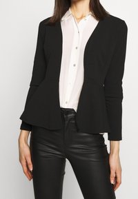 Žena v černém peplum saku přes bílou košili s knoflíky a černých kožených kalhotách, stojící na pozadí se světlým jednobarevným pozadím.