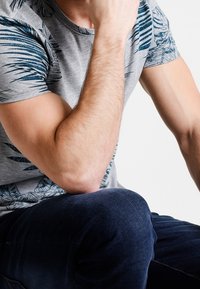 Grå t-shirt med blåt tropisk bladmønster på skuldrene. Model siddende, iført mørke jeans. Glat stof og afslappet pasform.