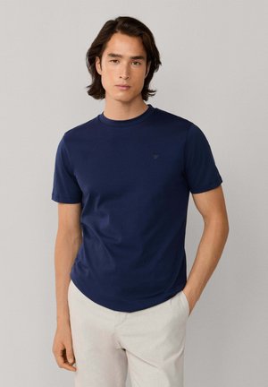 T-shirt à manches courtes bleu marine en matériau doux avec un col rond et un logo brodé subtil sur la poitrine, associé à un pantalon de couleur claire.