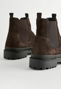 Bocanci Chelsea din suede maro, cu panouri elastice pe laterale, talpă groasă din cauciuc negru și tab-uri pentru o încălțare ușoară. Detalii cu o textură vizibilă.