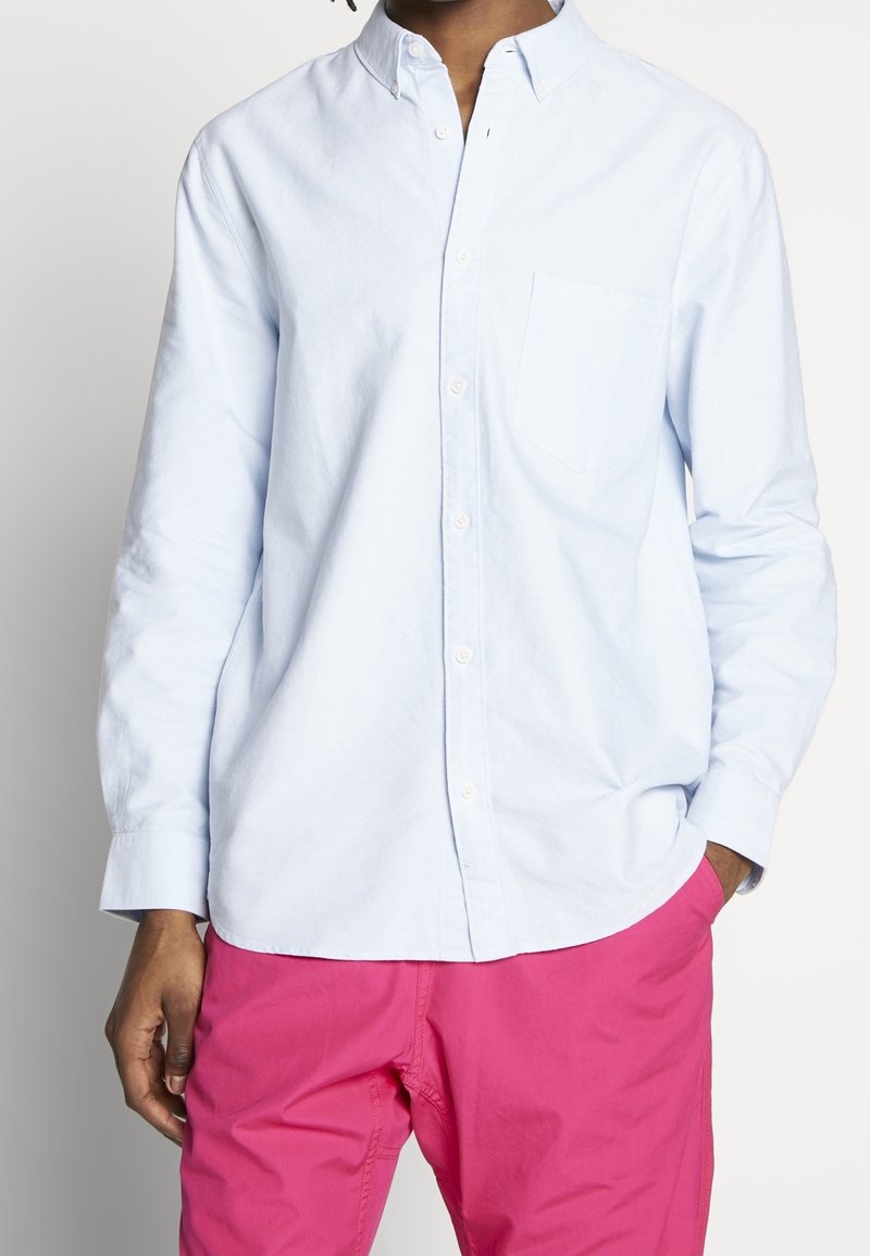 Chemise bleu clair en coton avec une poche poitrine unique, manches longues et col classique, rentrée dans un short rose.