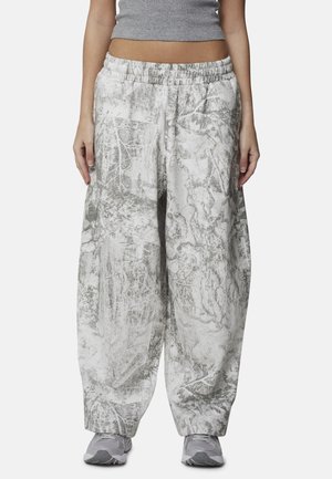 Personne portant un pantalon large gris et blanc à motif camouflage, des baskets grises et un crop top gris, debout devant un fond uni.