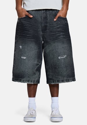 Persona que lleva pantalones cortos de mezclilla negros largos y holgados con detalles desgastados, calcetines blancos y zapatillas blancas, manos en los bolsillos.